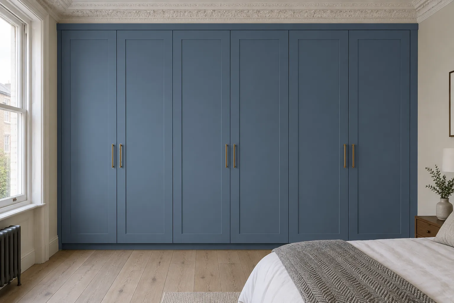 The same wardrobe painted a deep blue-grey against the pale walls and oak floor. The six Shaker-style doors keep their slim aged-brass bar handles, with the slim infill beneath the cornice, the side infills and the recessed plinth painted to match. This example shows how a deeper colour gives the wardrobe more presence in the room.