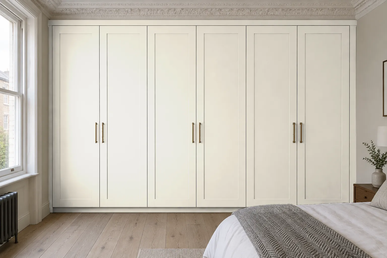 The same wardrobe painted a warm off-white close to the wall colour. The six Shaker-style doors keep their slim aged-brass bar handles, while the slim infill beneath the cornice, the side infills and the recessed plinth are painted to match. This example shows how a wardrobe painted in the wall colour blends into the room.